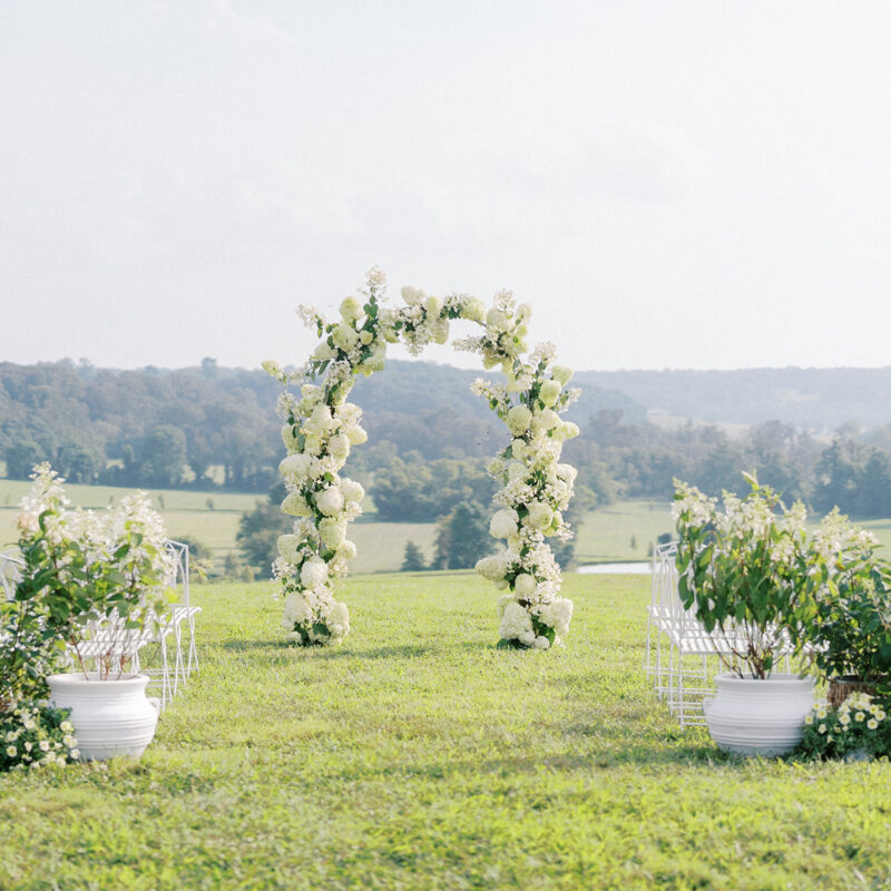 wedding ceremony in a green field with arch covered in white flowers and scenic views
