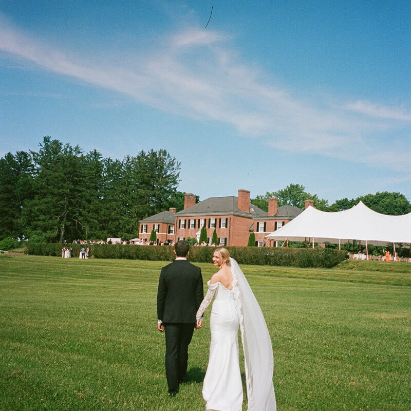 Bride and groom walking on grass towards Georgian estate with tented reception