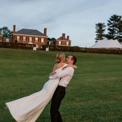 Groom carrying bride towards estate and tented reception