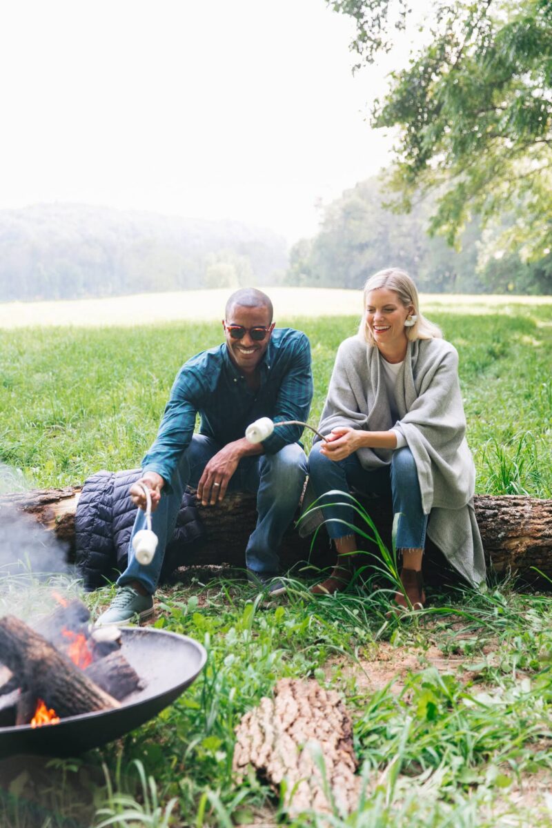 Two guests on a log toasting marshmallows over a fire pit at the edge of the meadow