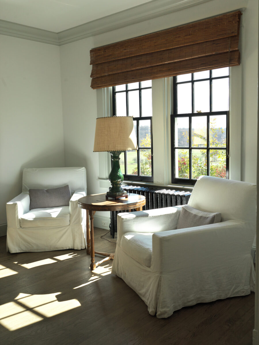 Two slipcovered armchairs in a sunlit corner of the guest house, at a window looking over the fields