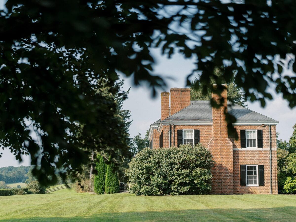 The brick manor with its slate roof and tall chimneys, framed by an oak canopy across the mown lawn