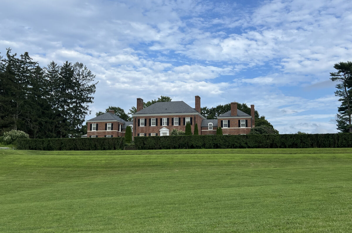 The Georgian manor house behind a clipped hedge, seen across the open lawn on a summer afternoon.