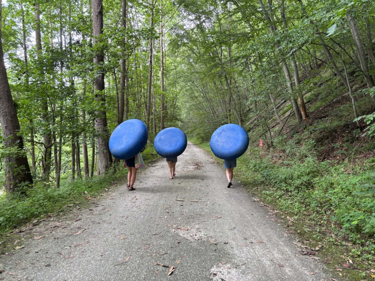 Three people carrying inner tubes down a shaded gravel lane through the woods