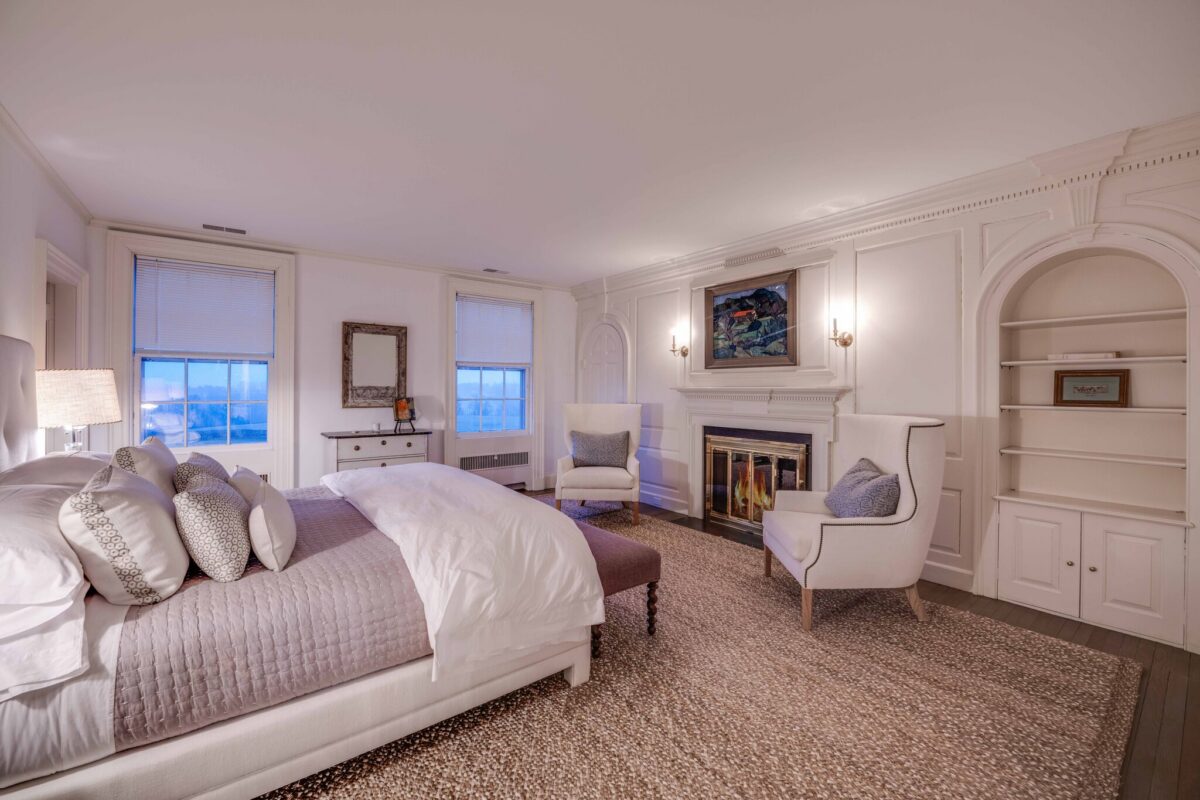 A bedroom in pale linens with a fire lit, arched built-in shelves and windows over the fields