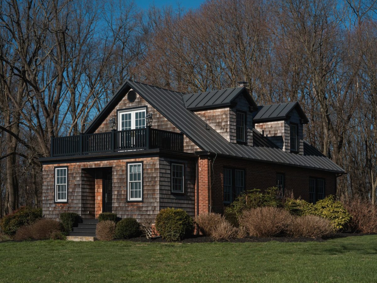 A cedar-shingled cottage with a standing-seam roof, dormer windows and an upper balcony, set against winter woods