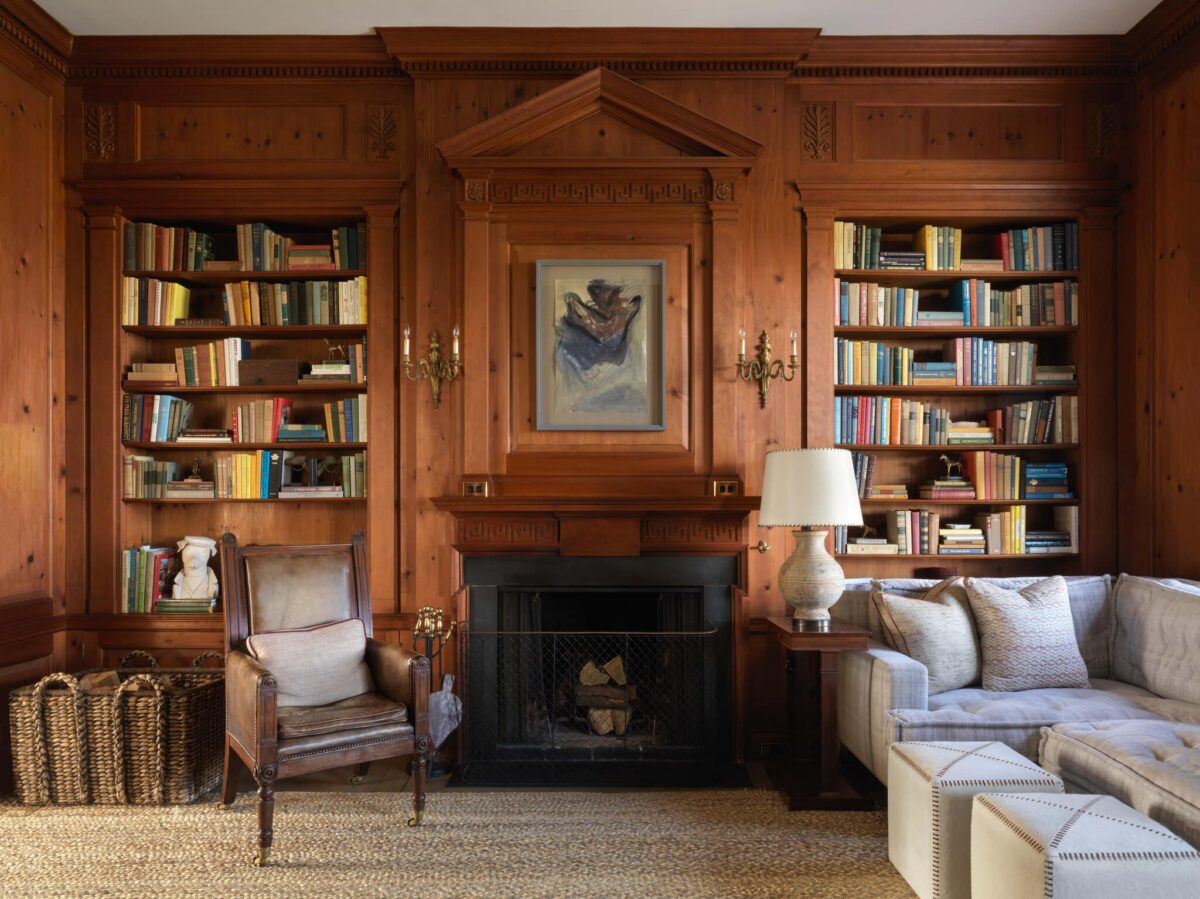 Pine-paneled library with floor-to-ceiling bookshelves, a carved mantel and a fire laid in the grate